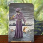 Tarot Card, the five of swords shows a robed person holding a sword in each hand, the aftermath of a battle in the background.